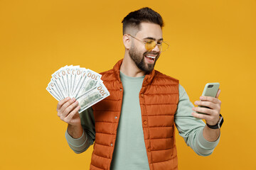 Wall Mural - Young smiling happy rich successful caucasian man in orange vest mint sweatshirt glasses holding mobile cell phone fan of cash money in dollar banknotes isolated on yellow background studio portrait