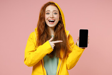 Redhead young woman in yellow waterproof raincoat hood outerwear point finger mobile cell phone blank screen workspace area isolated on pastel pink background studio Outdoor wet fall season concept