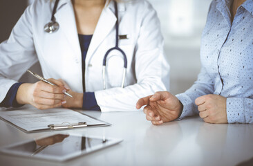 Wall Mural - Unknown woman- doctor is listening to her patient, while sitting together at the desk in the cabinet in a clinic. Female physician with a stethoscope is writing at clipboard, close up. Perfect medical