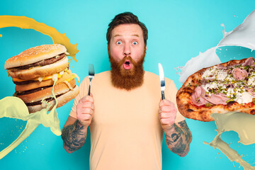 Wall Mural - Man with tattoos is ready to eat sandwich and pizza with cutlery in hand. cyan background