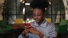 Crop view of afro american woman in blouse using smartphone while sitting in chair. Attractive female person smiling and browsing internet in cafe while spending fre time.