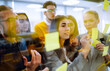 © maxbelchenko - Group of young modern people working on project together. Business colleagues writing on sticky note on glass wall with coworkers standing by in office. Startup Business. Planning, analysis.