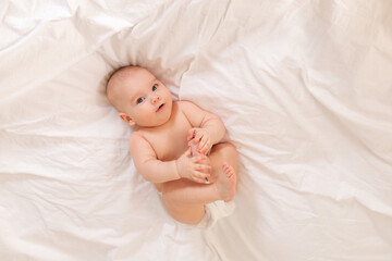 Cute baby in diapers lies in bed with white cotton linen, view from above, space for text