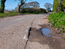 A Blue Farm Tractor Approaches Surface Damage To Tarmac On A Rural Road Is Marked For Repair With White Spray Paint. Taken On Wirral In The UK On A Sunny Day In Spring.