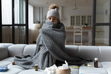 Sick Latin Female Feel Bad Ill Has Health Problems In Cold Freezing Flat With Broken Central Heating. Young Woman Sit On Sofa In Cap Covered With Knitted Plaid Taking Medicine For Fever Flu Influenza
