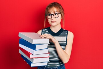 Sticker - Redhead young woman holding a pile of books smiling looking to the side and staring away thinking.