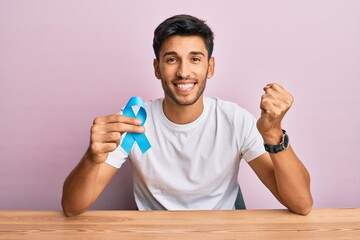 Young handsome man holding blue ribbon screaming proud, celebrating victory and success very excited with raised arm