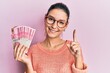 Young caucasian woman holding 100000 indonesian rupiah smiling with an idea or question pointing finger with happy face, number one