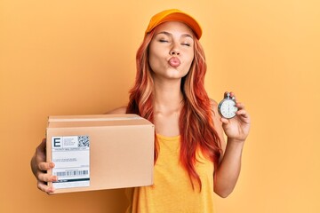 Wall Mural - Young redhead woman holding delivery package and stopwatch looking at the camera blowing a kiss being lovely and sexy. love expression.