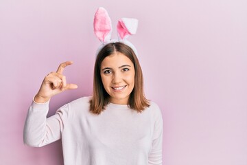 Canvas Print - Young beautiful woman wearing cute easter bunny ears smiling and confident gesturing with hand doing small size sign with fingers looking and the camera. measure concept.