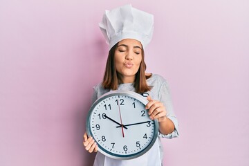 Canvas Print - Young beautiful woman wearing professional cook uniform and hat holding clock looking at the camera blowing a kiss being lovely and sexy. love expression.