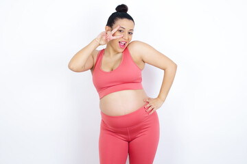 Sticker - young beautiful Arab pregnant woman in sports clothes against white wall making v-sign near eyes. Leisure, coquettish, celebration, and flirt concept.