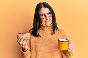 Poster - Middle age brunette woman eating pastry and drinking coffee in shock face, looking skeptical and sarcastic, surprised with open mouth