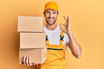 Wall Mural - Young handsome delivery man holding packages doing ok sign with fingers, smiling friendly gesturing excellent symbol