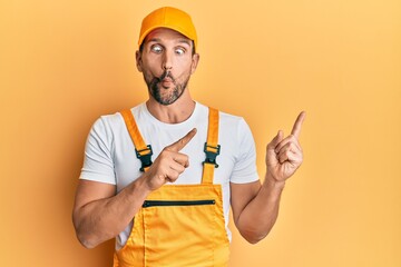 Canvas Print - Young handsome man wearing handyman uniform pointing to the side making fish face with mouth and squinting eyes, crazy and comical.