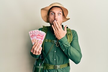 Canvas Print - Handsome man with beard wearing explorer hat holding 100 yuan banknotes covering mouth with hand, shocked and afraid for mistake. surprised expression