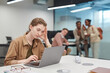 Portrait of stressed young woman using laptop in office or coworking space with group of people in background, copy space