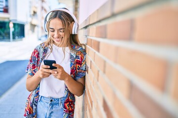 Wall Mural - Young beautiful blonde caucasian woman smiling happy outdoors on a sunny day wearing headphones and using smartphone leaning on a brick wall