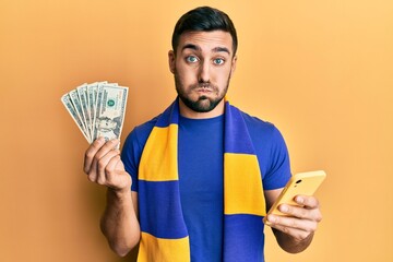 Poster - Young hispanic man football supporter using smartphone holding usa dollars banknotes puffing cheeks with funny face. mouth inflated with air, catching air.