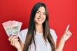 © Krakenimages.com - Young brunette woman holding 100 new zealand dollars banknotes smiling happy pointing with hand and finger to the side