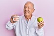 Senior caucasian man holding denture and green apple winking looking at the camera with sexy expression, cheerful and happy face.