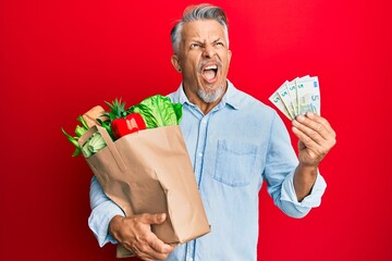 Poster - Middle age grey-haired man holding groceries and euro banknotes angry and mad screaming frustrated and furious, shouting with anger. rage and aggressive concept.