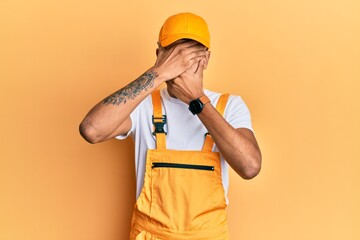 Canvas Print - Young handsome african american man wearing handyman uniform over yellow background covering eyes and mouth with hands, surprised and shocked. hiding emotion