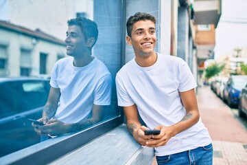 Poster - Young latin man smiling happy using smartphone leaning on the wall at the city.