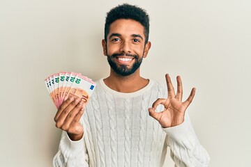 Wall Mural - Handsome hispanic man with beard holding bunch of 10 euro banknotes doing ok sign with fingers, smiling friendly gesturing excellent symbol