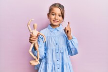 Little Beautiful Girl Holding Small Wooden Manikin Smiling With An Idea Or Question Pointing Finger With Happy Face, Number One