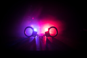 Police raid at night and you are under arrest concept. Silhouette of handcuffs with police car on backside. Image with the flashing red and blue police lights at foggy background.