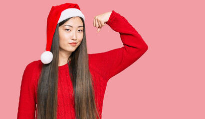 Wall Mural - Young chinese woman wearing christmas hat strong person showing arm muscle, confident and proud of power