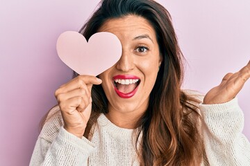 Sticker - Beautiful hispanic woman holding heart shape paper covering eye celebrating achievement with happy smile and winner expression with raised hand