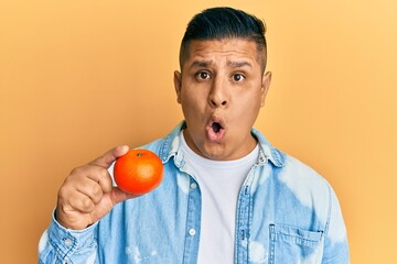 Canvas Print - Young latin man holding tangerine scared and amazed with open mouth for surprise, disbelief face