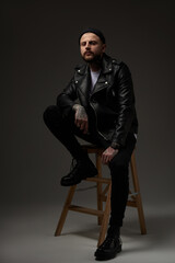 Invidivual and stylish guy with tattooed body and modern hairstyle in black suit poses in dark background looking at camera with serious face.