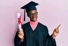 Young African American Girl Wearing Graduation Cap And Ceremony Robe Holding Diploma Smiling Happy Pointing With Hand And Finger To The Side