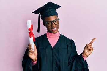 Wall Mural - Young african american girl wearing graduation cap and ceremony robe holding diploma smiling happy pointing with hand and finger to the side