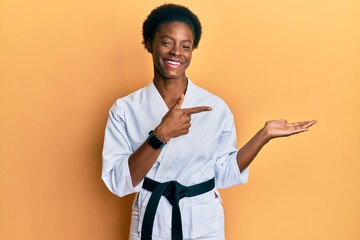 Canvas Print - Young african american girl wearing karate kimono and black belt amazed and smiling to the camera while presenting with hand and pointing with finger.