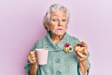 Wall Mural - Senior grey-haired woman drinking a cup of coffee and eating bun clueless and confused expression. doubt concept.