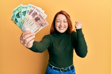 Poster - Beautiful redhead woman holding south korean won banknotes screaming proud, celebrating victory and success very excited with raised arm