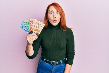 Sticker - Beautiful redhead woman holding canadian dollars scared and amazed with open mouth for surprise, disbelief face