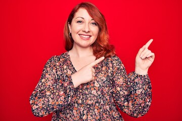 Canvas Print - Young beautiful redhead woman wearing floral casual dress standing over red background smiling and looking at the camera pointing with two hands and fingers to the side.