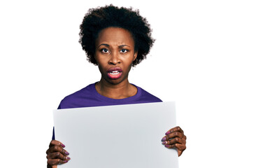 Sticker - African american woman with afro hair holding blank empty banner in shock face, looking skeptical and sarcastic, surprised with open mouth