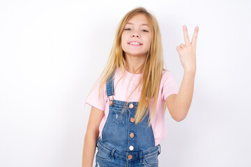 beautiful caucasian little girl wearing jeans overall over white background smiling and looking friendly, showing number two or second with hand forward, counting down