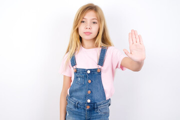 Wall Mural - beautiful caucasian little girl wearing jeans overall over white background shows stop sign prohibition symbol keeps palm forward to camera with strict expression