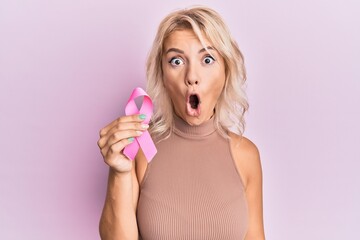 Poster - Young blonde girl holding pink cancer ribbon scared and amazed with open mouth for surprise, disbelief face