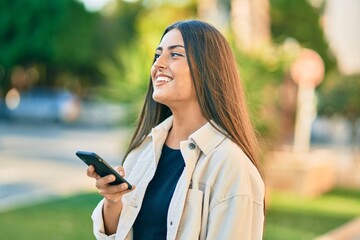 Wall Mural - Young hispanic girl smiling happy using smartphone at the park.