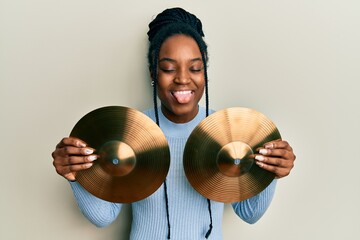 Wall Mural - African american woman with braided hair holding golden cymbal plates sticking tongue out happy with funny expression.