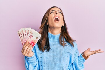 Poster - Young brunette woman holding 20 israel shekels banknotes crazy and mad shouting and yelling with aggressive expression and arms raised. frustration concept.