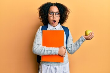 Wall Mural - Young little girl with afro hair wearing school bag holding books and green apple afraid and shocked with surprise and amazed expression, fear and excited face.